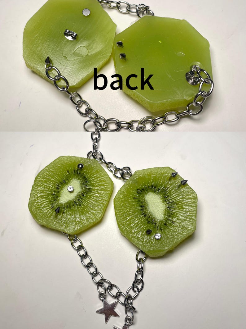 May include: A necklace featuring two green kiwi fruit slice pendants connected by a silver-toned metal chain. The pendants are embellished with small metal spikes and clear rhinestones. The word back is printed between the two pendants.