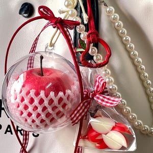 May include: A decorative bag charm featuring a clear spherical ornament containing a red apple wrapped in a white protective mesh, accented with a red gingham ribbon, a red apple-shaped metal charm, and a string of white faux pearls.