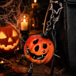 May include: A pumpkin-shaped keychain featuring a bright orange surface with a carved, smiling face, black accents, and silver-toned metal spikes protruding from the sides. A small black coffin-shaped charm hangs from the bottom of the pumpkin.
