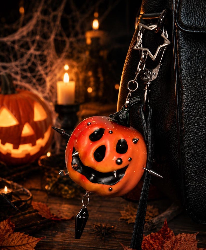 May include: A pumpkin-shaped keychain featuring a bright orange surface with a carved, smiling face, black accents, and silver-toned metal spikes protruding from the sides. A small black coffin-shaped charm hangs from the bottom of the pumpkin.