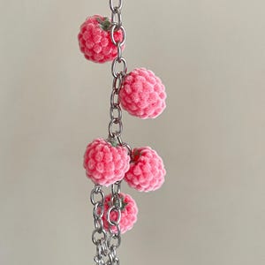 May include: A silver-toned metal chain keychain featuring five small, textured, pink raspberry-shaped charms with tiny green stems. The charms are attached at intervals along the vertical chain, which terminates in a small, pointed silver star charm.