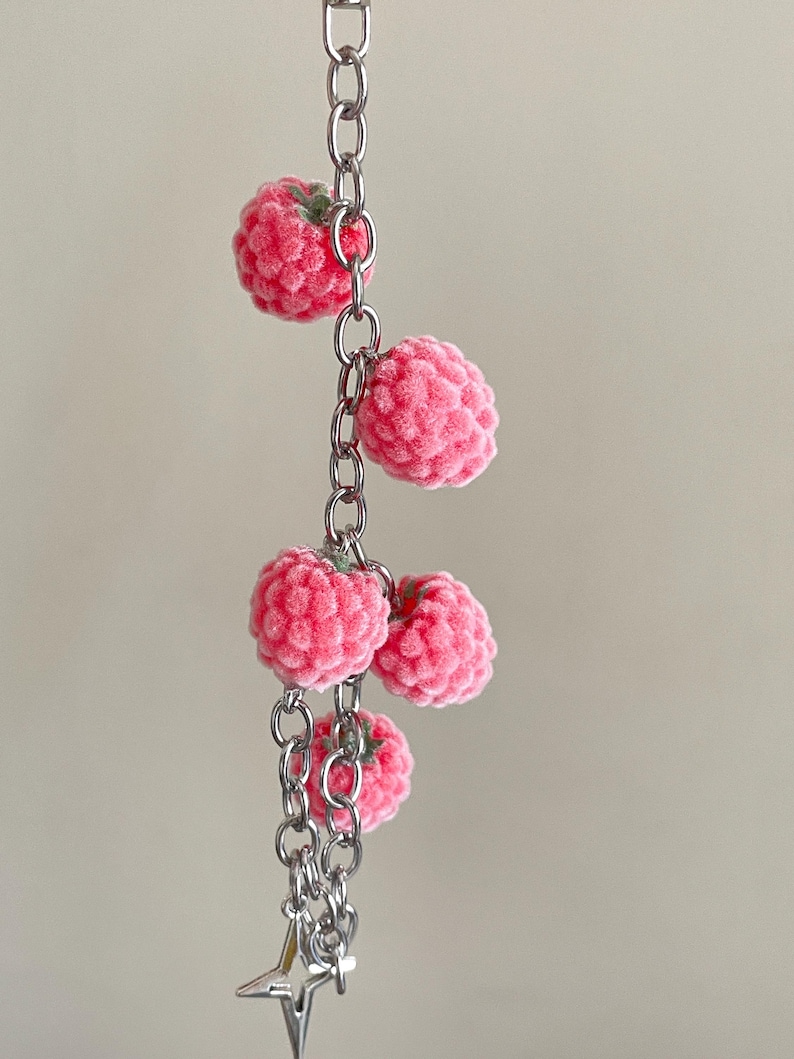 May include: A silver-toned metal chain keychain featuring five small, textured, pink raspberry-shaped charms with tiny green stems. The charms are attached at intervals along the vertical chain, which terminates in a small, pointed silver star charm.