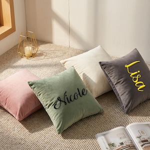 Pode incluir: Quatro almofadas quadradas de veludo em tons de rosa, verde, creme e cinza. A almofada verde tem "Nicole" bordado em preto, e a almofada cinza tem "Lisa" bordado em amarelo. Um castiçal geométrico dourado está no fundo.