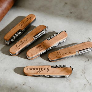 May include: Five wooden pocket knives with stainless steel tools and personalized names engraved on the wood. The names are in cursive script. The knives are arranged on a light gray surface, with a brown leather item in the background.