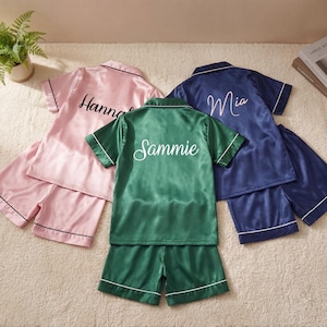 May include: Three sets of personalized satin pajamas in pink, green, and navy blue. Each set includes a short-sleeved top and shorts, with white trim. The names "Hannah", "Sammie", and "Mia" are written on the tops in cursive.