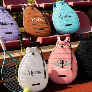 Op de afbeelding: Een verzameling gewatteerde tennisracket tassen in verschillende kleuren, waaronder paars, bruin, blauw, wit, roze en zwart. Elke tas heeft een trekkoordsluiting en een naam erop gedrukt. Een tennisracket en bal zijn ook zichtbaar.