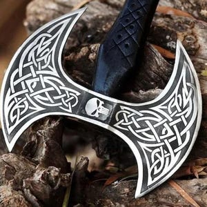 Könnte beinhalten: Eine Doppelklingen-Axt mit schwarzem Griff und silbernen Klingen. Die Klingen weisen komplizierte keltische Knotenmuster und ein Totenkopf-Emblem auf. Die Axt liegt auf einer verwitterten Holzoberfläche.