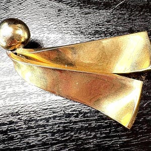 May include: A gold-toned metal brooch with a spherical top and two curved, ribbon-like elements. The brooch has a polished finish, reflecting light. The design is abstract and sculptural, suitable for adding a touch of elegance to any outfit.