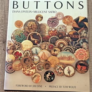 Puede incluir: Un libro de tapa dura titulado BUTTONS de Diana Epstein y Millicent Safro, con un prólogo de Jim Dine y un prefacio de Tom Wolfe. La cubierta blanca presenta un collage denso y colorido de varios botones decorativos con imágenes.
