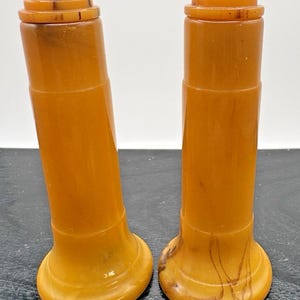 Pode incluir: Dois saleiros e pimenteiros cilíndricos de cor âmbar. Cada saleiro e pimenteiro tem uma base alargada e um topo em camadas. Os saleiros e pimenteiros são feitos de um material translúcido, com estrias e variações de cor visíveis.