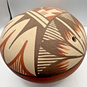 May include: A round, hand-painted ceramic vessel featuring a matte finish with geometric patterns in shades of terracotta, dark brown, and cream. The surface displays intricate line work and triangular motifs, with a small circular opening near the edge.