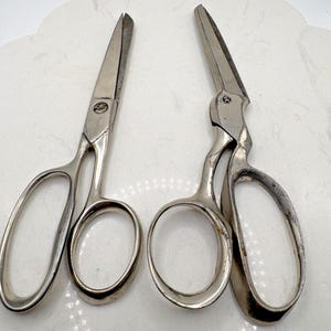 May include: Two pairs of silver-toned metal scissors with large oval finger loops. The left pair features a straight blade design, while the right pair has an offset handle and angled blades. Both tools show metallic surfaces with visible wear.