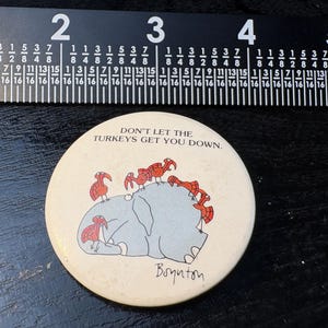 Op de afbeelding: Een ronde, gebroken witte knoop met de tekst "DON'T LET THE TURKEYS GET YOU DOWN." toont een cartoon olifant met kalkoenen op zijn rug. De knoop heeft een diameter van ongeveer 7,6 cm.