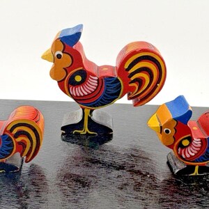 May include: Three hand-painted wooden rooster figurines in varying sizes. Each rooster features a vibrant color palette of red, blue, yellow, and black with stylized wing and tail feather patterns. The figures stand on black bases.