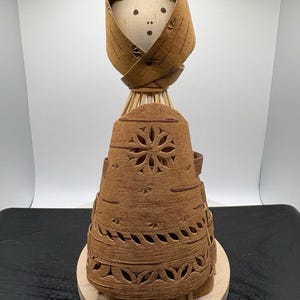 May include: A handcrafted figurine made from light brown birch bark, featuring a smooth wooden head with a simple painted face. The figure wears a bark hood and a flared, bell-shaped skirt decorated with intricate, laser-cut floral patterns.