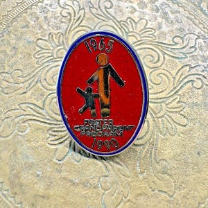 May include: Oval pin with a red background, blue border, and the text "1965" at the top. The pin features a stylized orange and black figure holding a smaller black figure, with "FOSTER GRANDPARENT PROGRAM 1990" at the bottom.