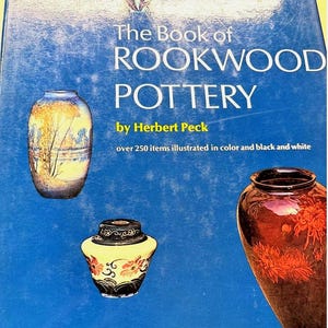 May include: A book cover titled "The Book of Rookwood Pottery" by Herbert Peck, featuring images of Rookwood pottery vases. The cover is blue with the title in white and the author's name in yellow. The book states it has over 250 items illustrated in color and black and white.