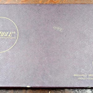 May include: A rectangular Scrabble game box with a textured, dark purple surface. The word "SCRABBLE" is in gold script inside a gold circle. The Selchow & Righter Co. logo is in gold on the lower right.