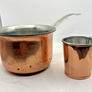 May include: A copper saucepan with a silver-toned interior and a long metal handle, positioned next to a smaller copper cup with a silver-toned interior and a small metal handle. Both items feature a polished metallic exterior finish.