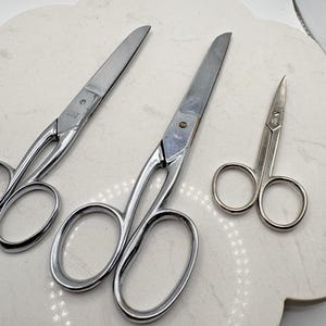 May include: Three silver-toned metal scissors of varying sizes arranged in a row. The two larger pairs feature elongated handles and pointed blades, while the smallest pair is a compact design with rounded finger loops and short, sharp blades.