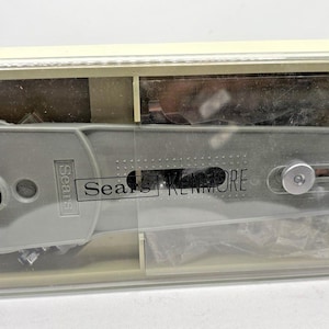 Könnte beinhalten: Ein grauer Sears Kenmore Nähmaschinenaufsatz in einem durchsichtigen Kunststoff-Aufbewahrungskoffer. Das Werkzeug hat eine längliche Form mit einem mittigen Schlitz, einer kreisförmigen Öffnung an einem Ende und einem silbernen Einstellknopf. Der Markenname Sears Kenmore ist in Schwarz auf der grauen Oberfläche aufgedruckt.