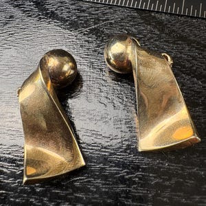 May include: A pair of gold-toned earrings. Each earring features a spherical top and a curved, rectangular body. The earrings are displayed on a dark, textured surface with a stainless steel ruler in the background.