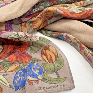 May include: A Liz Claiborne scarf featuring a floral print with orange, blue, and green flowers on a taupe background. The fabric has a soft, draped texture with a solid tan border section visible, showcasing a detailed botanical pattern throughout.