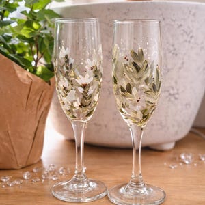 May include: Two hand-painted champagne flutes with floral designs. The clear glasses feature white flowers and green leaves. They sit on a wooden surface, with a plant and decorative items in the background.