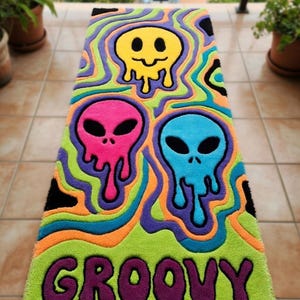 Könnte beinhalten: Ein rechteckiger Teppich mit einem lebendigen, psychedelischen Design. Er zeigt drei außerirdische Gesichter in Gelb, Pink und Blau mit dem Wort "GROOVY" in Lila. Der Teppich hat eine grüne Basis mit wellenförmigen Mustern in Orange, Lila und Blau.