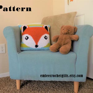 May include: A crocheted fox pillow with orange, white, and green stripes sits on a light blue armchair with a brown teddy bear. The armchair has wooden legs and is in a room with white walls. The text "emleecrochetgifts.etsy.com" is printed on the armchair.