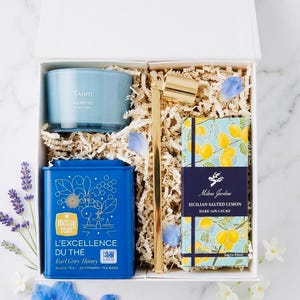 May include: A gift box with a blue candle, a blue tea tin, a gold-colored spoon, and a box of Sicilian salted lemon dark chocolate. The tea tin reads "L'EXCELLENCE DU THÉ Earl Grey Honey". The chocolate box has a lemon design.