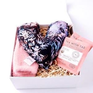 Puede incluir: Una caja de regalo blanca que contiene una almohada en forma de corazón con un estampado floral negro y azul, un artículo rosa difuso y dos paquetes de té rosas. La caja está llena de papel triturado. Los paquetes de té dicen "TEA WITH TAE".