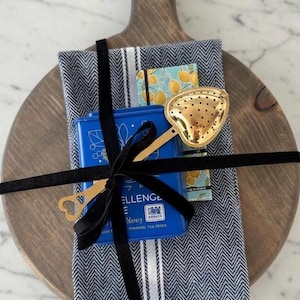 以下が含まれることがあります： 木製のまな板に乗ったギフトセット。青いお茶の缶、金色の茶こし、模様入りのティータオル、小さなレモンをテーマにしたカードが含まれています。黒いベルベットのリボンでまとめられています。