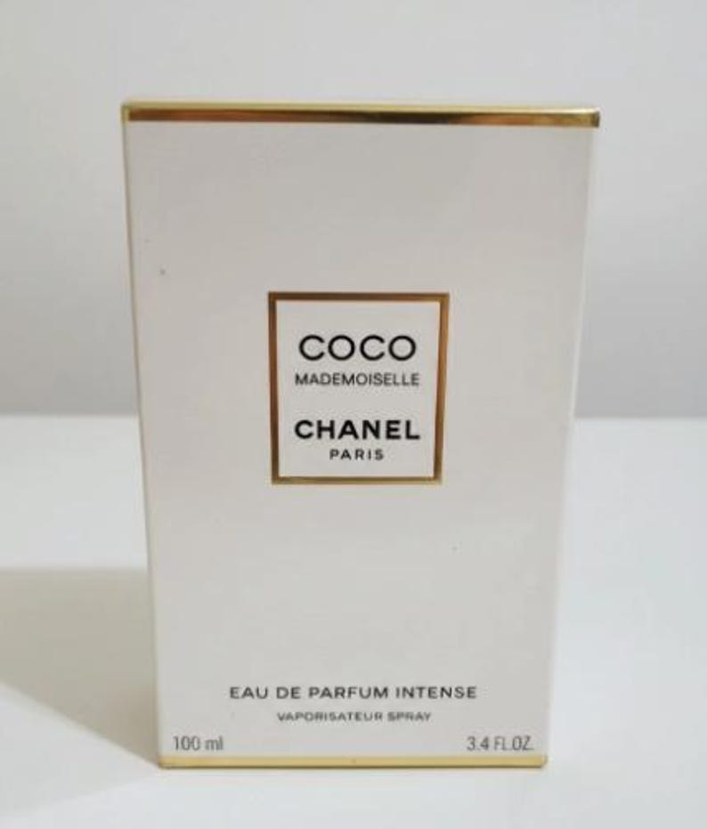 May include: White rectangular box with gold trim. The box features the text "COCO MADEMOISELLE CHANEL PARIS" in a gold-bordered rectangle. Below, it reads "EAU DE PARFUM INTENSE VAPORISATEUR SPRAY". The box also indicates 100 ml and 3.4 FL.OZ.