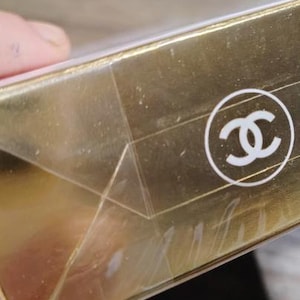 May include: Close-up of a gold-colored box with a white logo. The box has a clear, plastic wrap. The logo is a white, interlocking double-C design inside a white circle. The box appears to be made of cardboard.