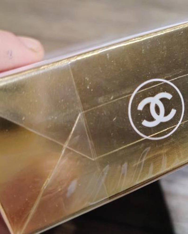 May include: Close-up of a gold-colored box with a white logo. The box has a clear, plastic wrap. The logo is a white, interlocking double-C design inside a white circle. The box appears to be made of cardboard.
