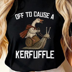 Könnte beinhalten: Schwarzes T-Shirt mit dem Text "OFF TO CAUSE A KERFUFFLE". Die Grafik zeigt eine Gans mit Umhang, die eine Schnecke reitet. Die Gans ist weiß mit rotem Umhang, die Schnecke braun und grau.