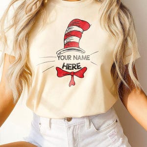 Op de afbeelding: Crèmekleurig T-shirt met een rood-wit gestreept hoedontwerp, een rode vlinderdas en de tekst "YOUR NAME HERE" in grijs. Het ontwerp is een speelse en grillige afbeelding.