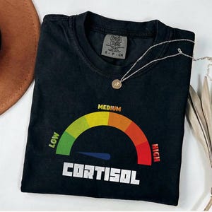 Könnte beinhalten: Schwarzes T-Shirt mit einer Grafik einer Anzeige, die "Cortisol" anzeigt. Die Anzeige reicht von "Low" bis "High", wobei der Zeiger auf "Medium" zeigt. Eine silberne Halskette mit einem kleinen Anhänger liegt auf dem T-Shirt.