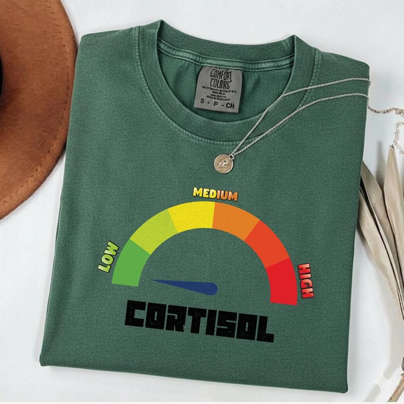 Puede incluir: Camiseta verde con un gr&aacute;fico de medidor de cortisol, que muestra un rango de "BAJO" a "ALTO". La palabra "CORTISOL" est&aacute; impresa debajo. Un collar plateado con un colgante est&aacute; sobre la camiseta.