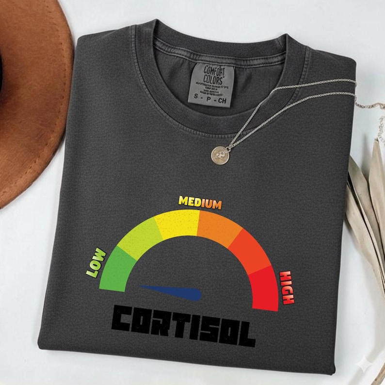 Puede incluir: Camiseta gris oscuro Comfort Colors con un gr&aacute;fico de medidor de cortisol. El medidor muestra un rango de verde (bajo) a rojo (alto), con la palabra "CORTISOL" debajo. Tambi&eacute;n es visible un collar plateado con un colgante.