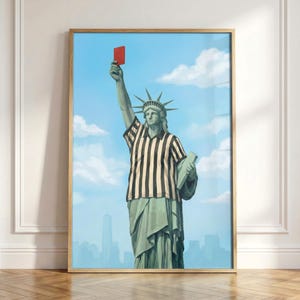 May include: Framed art print featuring the Statue of Liberty in a referee's uniform, holding a red card aloft. The artwork depicts the statue against a blue sky with clouds and a cityscape in the background. The print is in a wooden frame.
