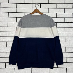 May include: A color-blocked sweatshirt in gray, white, and navy blue. The top section is gray, followed by a white band, and the lower portion is navy. The sweatshirt is hanging on a wooden hanger against a white brick wall.