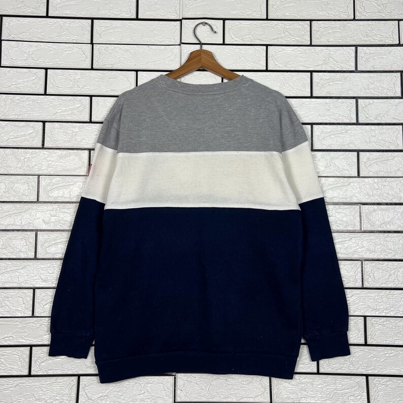 May include: A color-blocked sweatshirt in gray, white, and navy blue. The top section is gray, followed by a white band, and the lower portion is navy. The sweatshirt is hanging on a wooden hanger against a white brick wall.