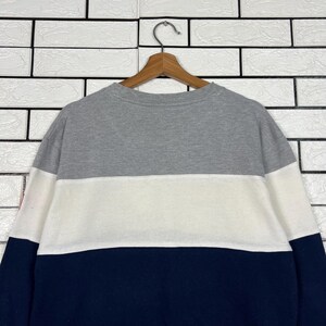 May include: A color-block sweatshirt in gray, white, and navy blue, hanging on a wooden hanger. The sweatshirt has a crew neck and long sleeves. The color-block design features a gray upper section, a white middle section, and a navy blue lower section.