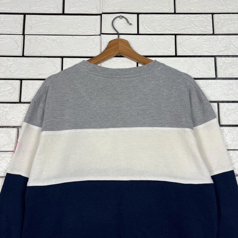 May include: A color-block sweatshirt in gray, white, and navy blue, hanging on a wooden hanger. The sweatshirt has a crew neck and long sleeves. The color-block design features a gray upper section, a white middle section, and a navy blue lower section.