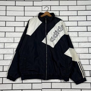 Può includere: Giacca da tuta Adidas nera e bianca con chiusura a zip completa. La giacca presenta un design diagonale bianco sul petto con il logo Adidas in bianco. Le maniche hanno strisce bianche. La giacca è appesa a una gruccia di legno.