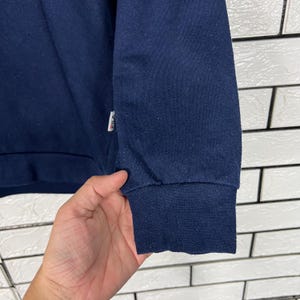 May include: Close-up of a navy blue sweatshirt sleeve. The cuff is ribbed and darker in color. A small white tag with red and blue accents is visible near the shoulder. The background is a white brick wall.