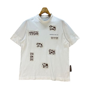 May include: White short-sleeved t-shirt with a mock neck. The shirt features brown and white cow illustrations on patches. The text "SPORTING GEAR KAI" is printed on the front. A brown tag is attached to the side.