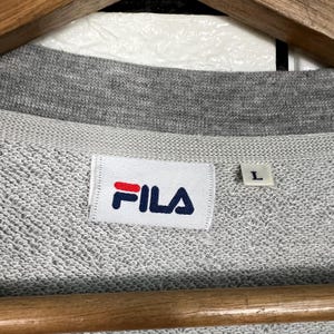 May include: Close-up of a gray FILA sweatshirt. The white label with the FILA logo in red and navy blue is visible. The size tag shows a size "L". The sweatshirt is on a wooden hanger.